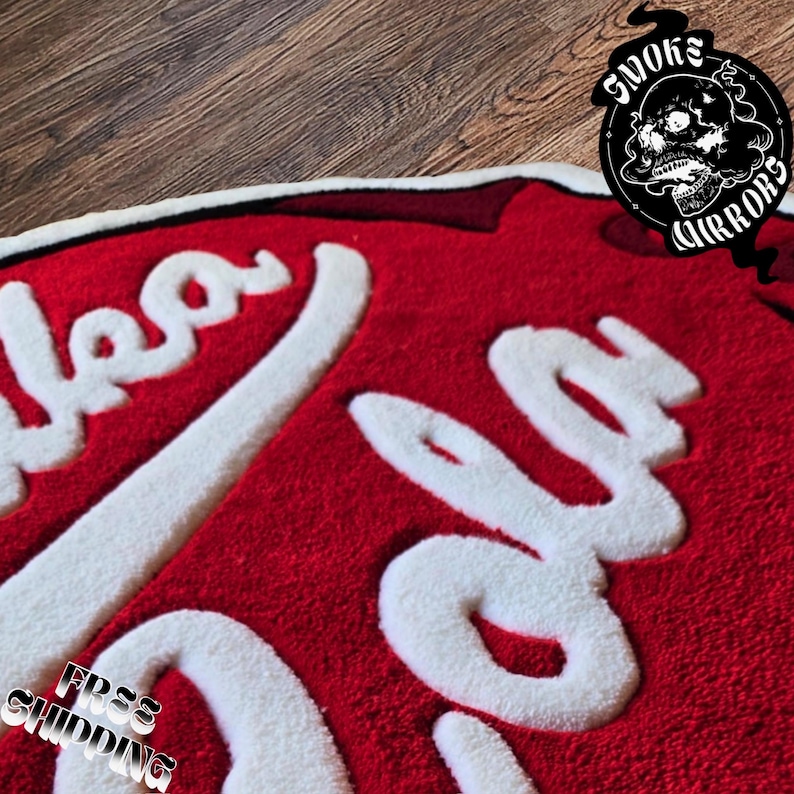 May include: Red and white rug with the word "cola" in a stylized font. The rug has a thick, textured pile and a white border. A black and white logo with a skull is in the upper right corner.