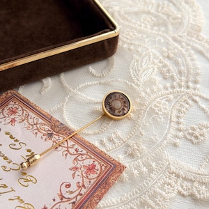 May include: A gold-toned lapel pin featuring a circular brown center with the text HERMES PARIS embossed in gold. The pin has a long, thin gold-toned metal stem and a small, rounded gold-toned end cap, presenting a classic and refined accessory design.