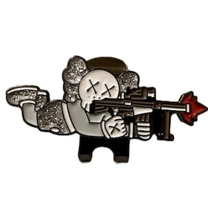 May include: A cartoon-style pin featuring a character with a skull-like face and crossed-out eyes, holding a rifle. The character is dressed in gray and black, with glitter accents on the arms and head. The rifle has a red muzzle flash.