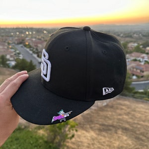 May include: Black baseball cap with the San Diego Padres logo in white. The cap features a small pin of a cartoon character. The New Era logo is visible on the side. The background shows a sunset over a residential area.