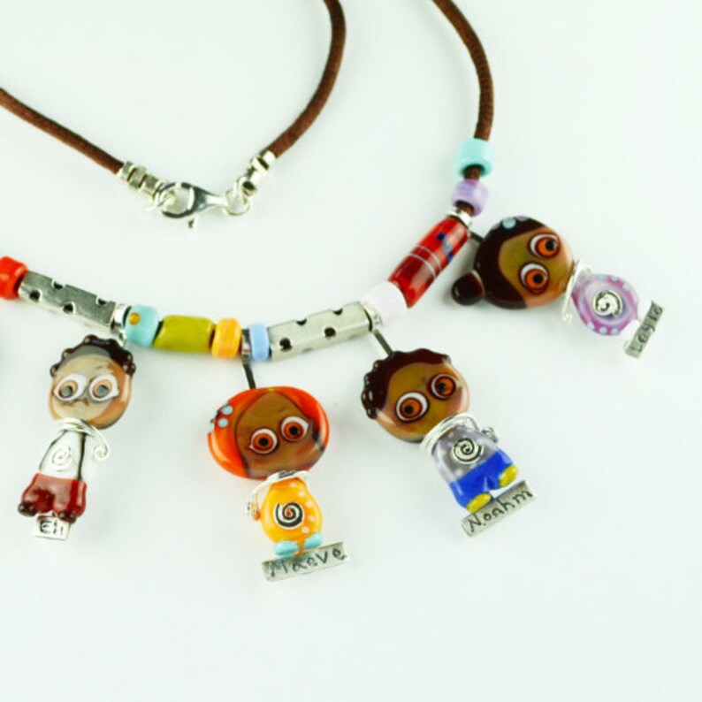 May include: A colorful necklace with five glass beads shaped like people, each with a different name tag. The beads are red, orange, yellow, green, blue, and purple. The necklace is made of brown leather cord and silver metal beads.