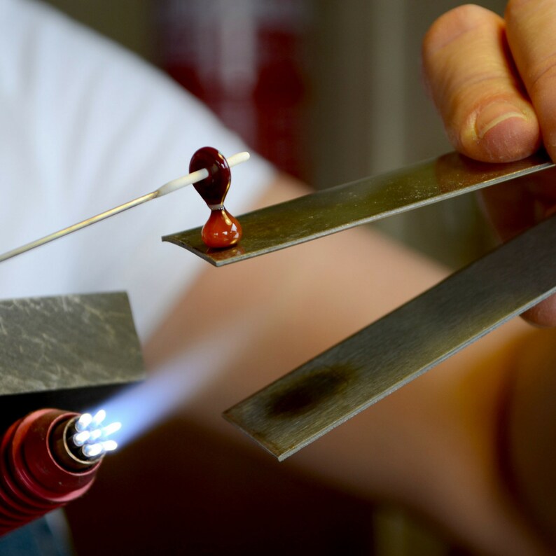 May include: A glass bead being heated with a torch while held by metal tongs. The bead is red and white and is being worked on by a glassblower.