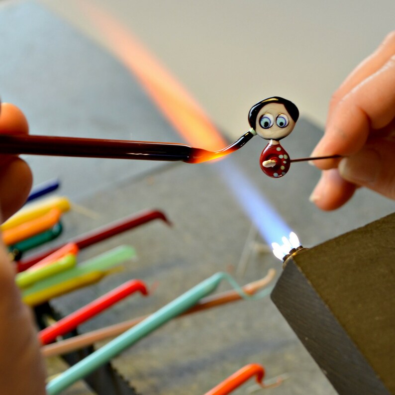 May include: A glass figurine of a person with black hair, a red dress with white polka dots, and blue eyes is being heated with a torch. The figurine is on a metal rod and is being held by a person's hand.
