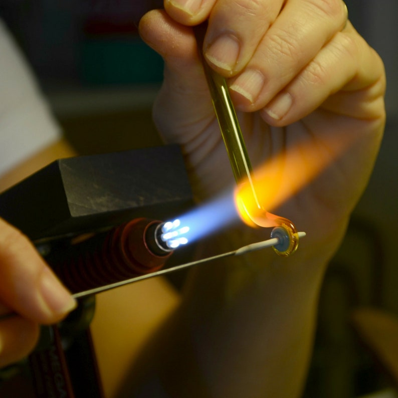 May include: A person is using a torch to heat a glass rod. The rod is being held over a metal stand with a blue flame coming from the torch. The glass is starting to melt and become more pliable.