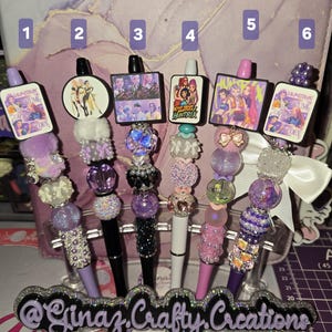 May include: A collection of six decorative pens with various designs. Each pen features a beaded body in shades of purple, with a charm at the top. The charms include images of groups and the text "HUNTR". The pens are displayed on a clear stand with the text "@Ginaz.Crafty.Creations".