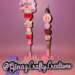 May include: Two decorative pens with a pink and strawberry theme. One pen features a Strawberry Shortcake character, while the other has a similar design. The pens are adorned with various beads and charms, including stars, hearts, and pom-poms. The base has the text "@Ginaz.Crafty.Creations".