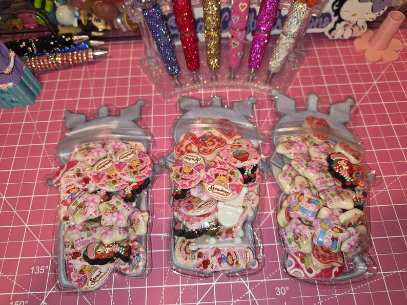 Puede incluir: Tres bolsas de pl&aacute;stico transparente con forma de personajes de dibujos animados, cada una llena de un surtido de peque&ntilde;os dijes de resina planos, circulares y en forma de estrella con coloridas ilustraciones de una ni&ntilde;a con sombrero rojo.