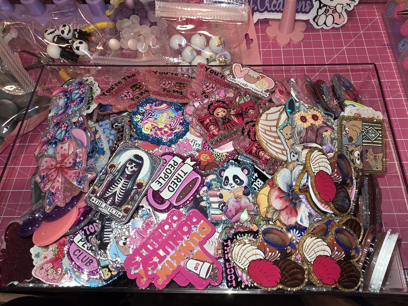 Puede incluir: Un contenedor de pl&aacute;stico transparente lleno de una variedad de dijes acr&iacute;licos coloridos y adornos planos. La colecci&oacute;n incluye dise&ntilde;os con esqueletos, pandas, flores, tazas de caf&eacute; y varios personajes de dibujos animados con acabados brillantes.