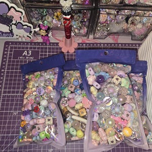 以下が含まれることがあります： 紫、ピンク、青、白の色合いのカラフルなクラフトビーズが詰まった3つの透明なプラスチックポーチ。ビーズには星、ハート、球体、花などの多様な形があり、一部には虹色やキラキラした仕上げが施されています。