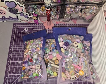 May include: Three clear plastic pouches filled with a variety of colourful craft beads in shades of purple, pink, blue, and white. The beads feature diverse shapes including stars, hearts, spheres, and flowers, with some having iridescent or glittery finishes.