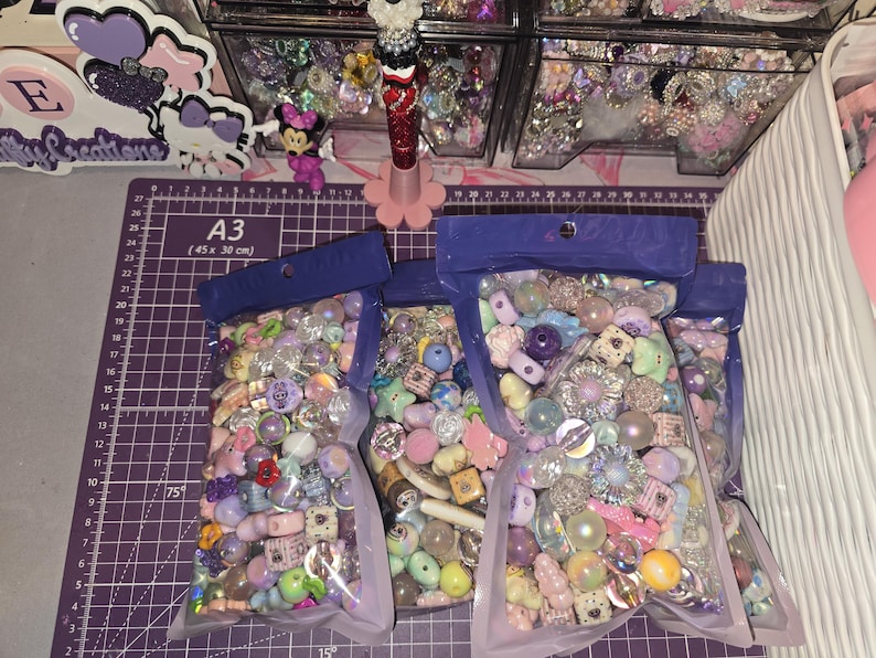 Puede incluir: Tres bolsas de pl&aacute;stico transparente llenas de una variedad de cuentas artesanales coloridas en tonos morado, rosa, azul y blanco. Las cuentas presentan diversas formas, incluyendo estrellas, corazones, esferas y flores, con acabados iridiscentes.