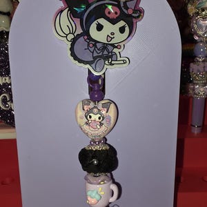 Puede incluir: Un portacredenciales con un adorno holográfico del personaje Kuromi, una cuenta en forma de corazón con una ilustración de Kuromi, una cuenta negra texturizada y un dije de taza color lavanda. El conjunto está montado en una tarjeta de color violeta claro con el texto Gina's Crafty Creations.