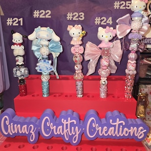 Puede incluir: Cinco bolígrafos decorativos numerados del #21 al #25, con figuras como Hello Kitty y Cinnamoroll, cuentas de colores y lazos. Los bolígrafos se exhiben en un soporte rojo de varios niveles con un letrero morado Cinaz Crafty Creations.