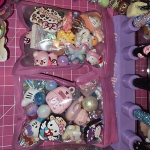 Puede incluir: Dos bolsas de pl&aacute;stico transparente llenas de cuentas y dijes artesanales variados, incluyendo figuras de Hello Kitty, un dije de mochila rosa, esferas iridiscentes y formas de mariposa sobre una base de corte rosa.