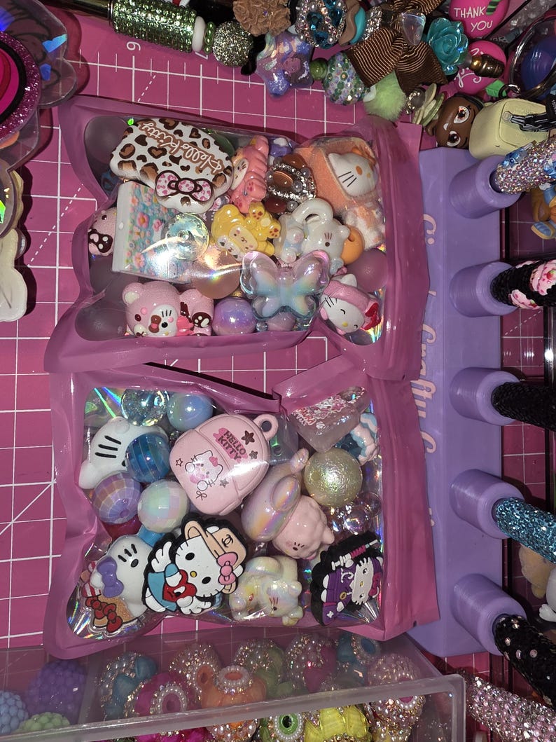 Puede incluir: Dos bolsas de pl&aacute;stico transparente llenas de cuentas y dijes artesanales variados, incluyendo figuras de Hello Kitty, un dije de mochila rosa, esferas iridiscentes y formas de mariposa sobre una base de corte rosa.