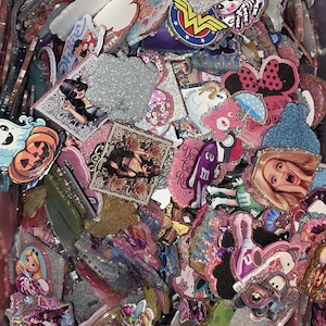 Puede incluir: Una colecci&oacute;n de dijes de acr&iacute;lico variados con diversos estampados gr&aacute;ficos coloridos, que incluyen personajes de dibujos animados, calaveras e &iacute;conos de la cultura pop. Muchas piezas tienen acabados brillantes, bordes texturizados y formas diversas.