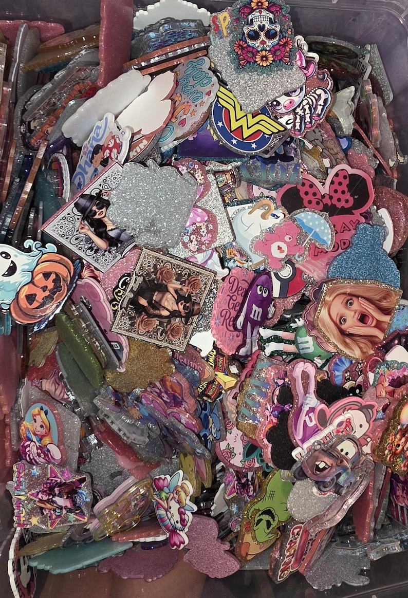 Puede incluir: Una colecci&oacute;n de dijes de acr&iacute;lico variados con diversos estampados gr&aacute;ficos coloridos, que incluyen personajes de dibujos animados, calaveras e &iacute;conos de la cultura pop. Muchas piezas tienen acabados brillantes, bordes texturizados y formas diversas.