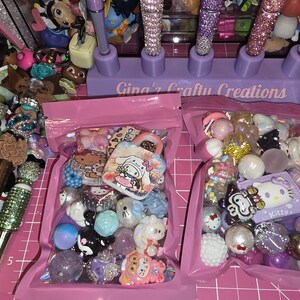Puede incluir: Dos bolsas de pl&aacute;stico rosa llenas de cuentas artesanales coloridas y dijes con temas de personajes, incluyendo dise&ntilde;os de Hello Kitty y Cinnamoroll. Un soporte morado con la etiqueta Gina's Crafty Creations sostiene bol&iacute;grafos brillantes.
