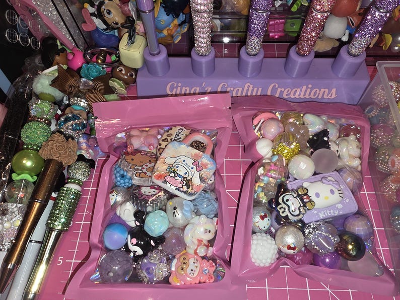 Puede incluir: Dos bolsas de pl&aacute;stico rosa llenas de cuentas artesanales coloridas y dijes con temas de personajes, incluyendo dise&ntilde;os de Hello Kitty y Cinnamoroll. Un soporte morado con la etiqueta Gina's Crafty Creations sostiene bol&iacute;grafos brillantes.