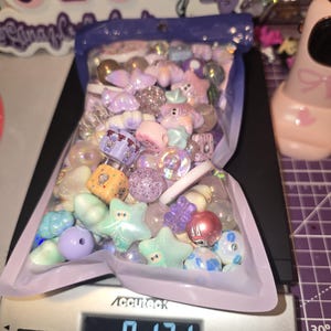 Puede incluir: Una bolsa de pl&aacute;stico transparente llena de una variedad de cuentas para manualidades con formas de estrellas, cubos y esferas, sobre una b&aacute;scula digital que muestra un peso de 343 gramos, con tonos pastel morados, azules y rosas.