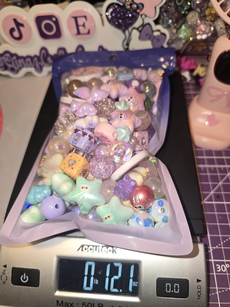 Puede incluir: Una bolsa de pl&aacute;stico transparente llena de una variedad de cuentas para manualidades con formas de estrellas, cubos y esferas, sobre una b&aacute;scula digital que muestra un peso de 343 gramos, con tonos pastel morados, azules y rosas.