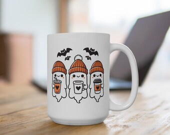 May include: White ceramic mug with a design of three ghosts wearing orange and white knit hats and holding cups of coffee. The ghosts are smiling and have black eyes. There are two black bats above the ghosts.