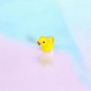 May include: A small, bright yellow rubber duck with a red beak and black eye. The duck is positioned on a reflective surface, with a soft gradient background of blue and pink hues. The toy is a classic bath time accessory.