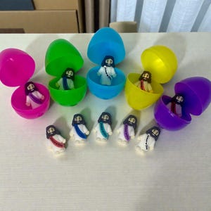 May include: A collection of colorful plastic Easter eggs, each opened to reveal a small figurine of Jesus. The figurines wear white robes and colored sashes with text. The eggs are in shades of pink, green, blue, yellow, and purple.