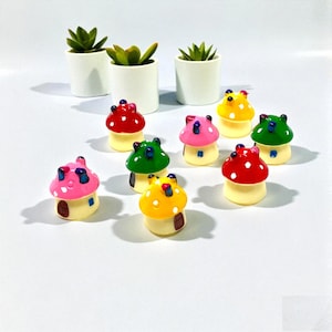 May include: A collection of colorful mushroom-shaped figurines with white polka dots on their caps. The figurines are red, pink, yellow, and green. Three small potted succulents are in the background, adding a touch of greenery.