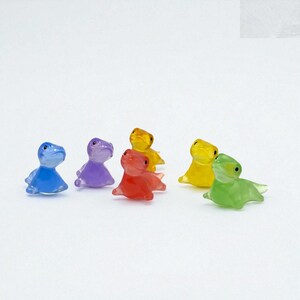 May include: A collection of six small, translucent dinosaur figurines in various colors: blue, purple, red, yellow, and green. Each figurine is posed in a sitting position, with a smooth, glossy finish. The toys are arranged on a white background.