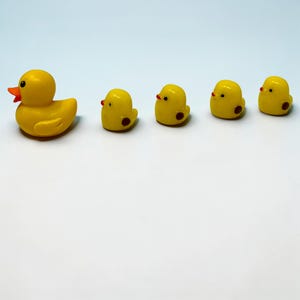 May include: A collection of yellow duck figurines. One larger duck with an orange beak is positioned to the left, with four smaller ducklings to the right. Each duckling has a red dot and a red beak. The figurines are on a white background.