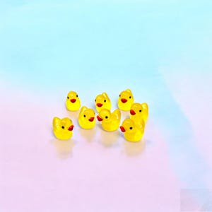 May include: A collection of eight small, yellow duck figurines with red beaks and black eyes. The ducks are arranged on a gradient background of pink and blue. These miniature ducks are likely decorative items.