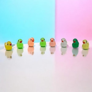 May include: A collection of eight miniature duck figurines in various colors, including yellow, green, orange, pink, and white. Each duck has a small orange beak and black eyes. The ducks are arranged in a row against a pastel background.