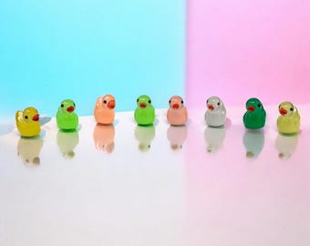 May include: A collection of eight miniature duck figurines in various colors, including yellow, green, orange, pink, and white. Each duck has a small orange beak and black eyes. The ducks are arranged in a row against a pastel background.