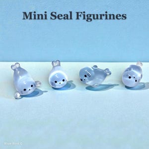 Può includere: Quattro piccole statuette di foca in azzurro traslucido con corpi lisci e arrotondati. Ogni foca presenta occhi dipinti di nero, un piccolo naso nero e baffi. Il testo 'Mini Seal Figurines' è in alto, 'Blue Bird Q' è in basso.