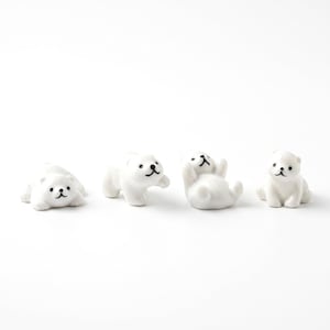 May include: Four small white ceramic bear figurines arranged in a row. Each bear has a smooth, glossy finish with simple black painted details for the eyes, nose, and mouth. The bears are depicted in different poses, including lying down and sitting.