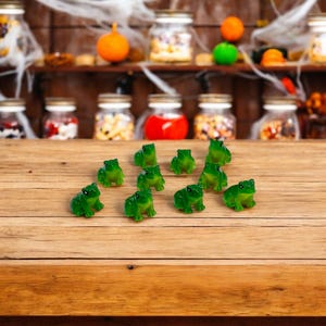 May include: A collection of green frog-shaped candies arranged on a wooden surface. The candies are translucent and appear to be gummy. The background features a shelf with jars of colorful treats, suggesting a Halloween theme.