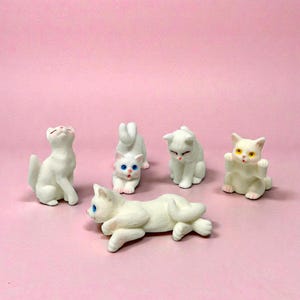 May include: A collection of white cat figurines in various poses, including sitting, stretching, and lying down. Some cats have blue or yellow eyes, set against a pink background. These decorative cat figurines are ideal for cat lovers.
