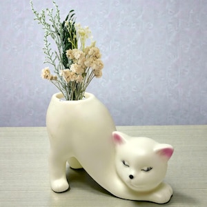 May include: A cream-colored cat-shaped ceramic vase with dried flowers. The vase is in the shape of a stretching cat, with pink accents on the ears. The vase holds a bouquet of dried flowers and greenery.