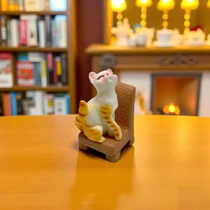 May include: A miniature figurine of a white and orange tabby cat sitting on a small brown chair. The cat is looking upwards with its eyes closed. The chair has a woven back and a square seat. The background includes a bookshelf and a fireplace.