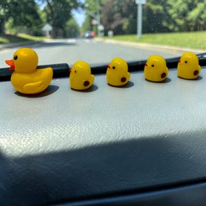 May include: A collection of yellow rubber duck and chick figurines. The duck has an orange beak, and the chicks have red beaks and brown spots. The figurines are arranged on a gray surface, with a blurred background of a road and trees.