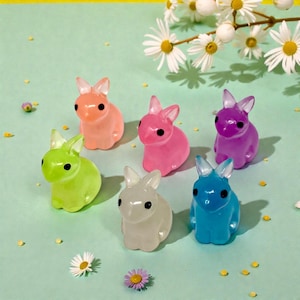 May include: Six colorful, translucent bunny figurines in shades of orange, pink, green, white, purple, and blue. Each bunny has black eyes and upright ears. The bunnies are arranged on a light green surface with flowers.