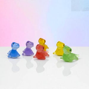 May include: A collection of seven small, translucent dinosaur figurines in various colors: blue, purple, red, yellow, and green. The figurines are posed in a sitting position, with a smooth, glossy finish. The background is a gradient of pink and blue.