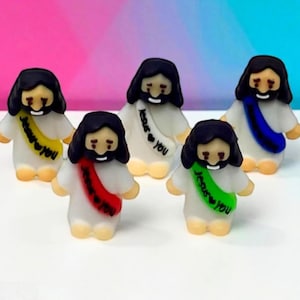 May include: Five small figurines of Jesus with black hair and beards, wearing white robes and colorful sashes. Each sash has the text "Jesus ❤️ You" in black. The background is a gradient of pink and blue.
