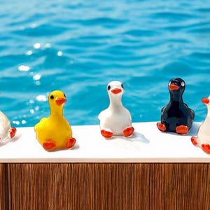 May include: Five ceramic duck figurines in white, yellow, and black with red beaks and feet. The ducks are sitting on a white surface with a blue water background. The ducks are a decorative item.