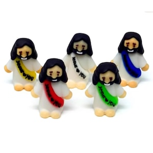 May include: Five small figurines of a person with long dark hair and a beard, wearing a white robe with a colored sash. Each sash is a different color—yellow, white, red, green, and blue—and features the printed text Jesus loves you.
