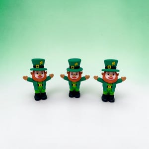 May include: Three small leprechaun figurines. Each figure wears a green hat with a shamrock, a green jacket, and black shoes. They have orange beards and are posed with their arms outstretched. The background is a gradient of light green.