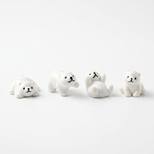 May include: Four small white ceramic bear figurines arranged in a row. Each bear has a smooth, glossy finish with simple black painted details for the eyes, nose, and mouth. The bears are depicted in different poses, including lying down and sitting.