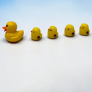 May include: A yellow rubber duck with an orange beak and four small yellow ducklings with red dots and beaks. The ducklings are lined up behind the larger duck. The background is a gradient of white to light blue.