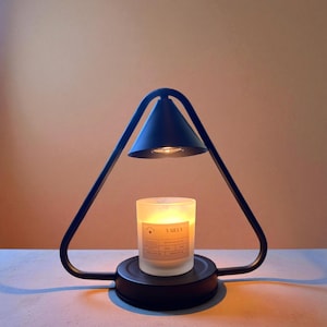 May include: A black candle warmer with a triangular frame and a conical shade. A vanilla-scented candle sits on the base, with a label visible. The warmer is designed to gently melt the candle, releasing its fragrance.